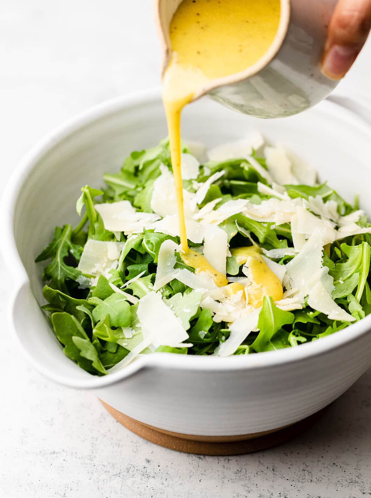 Hand pouring dressing over fresh arugula and parmesan cheese in a large white bowl.