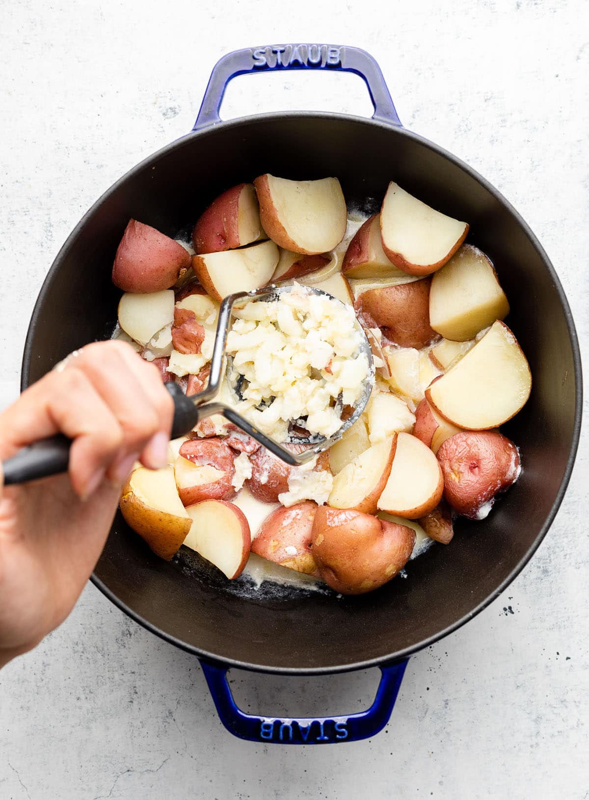 Hand using a potato masher to mash potatoes in a large black pot.