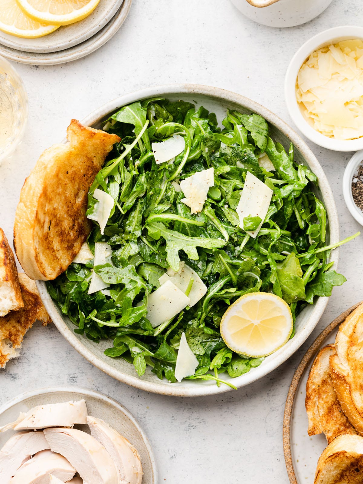 Overhead view of arugula salad in a white bowl, with toasted bread and lemon slices on the side.