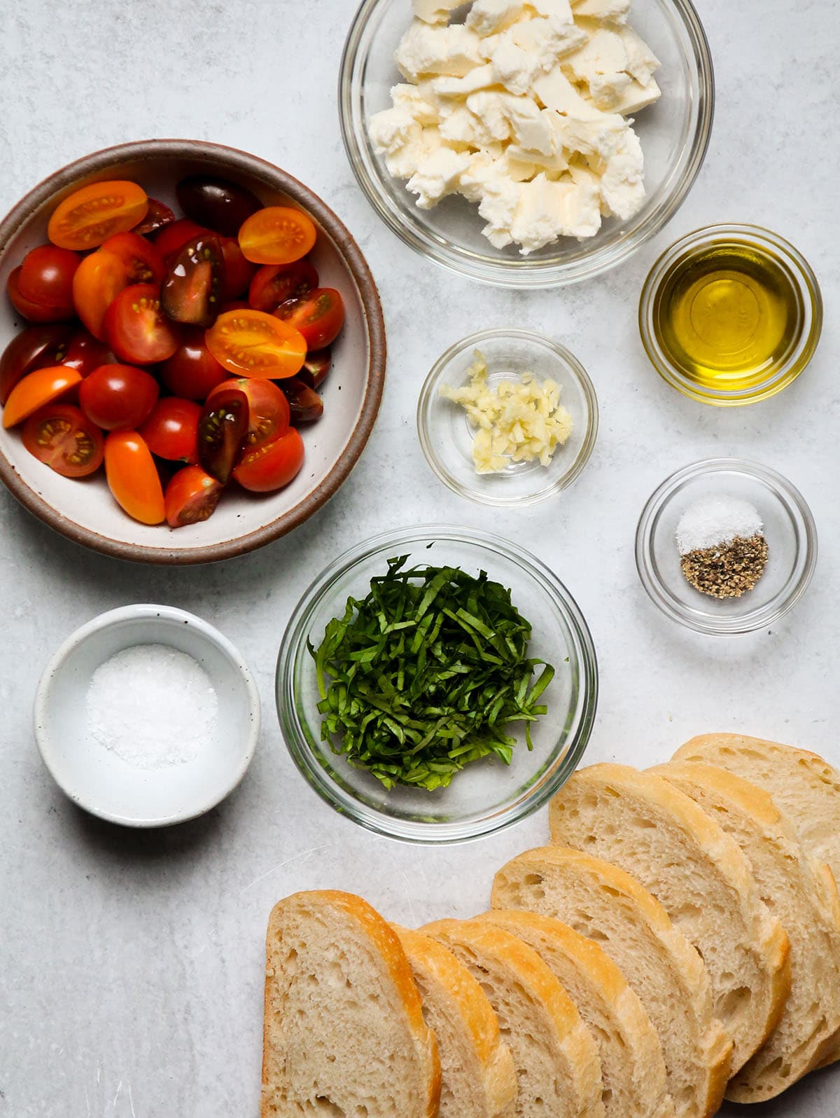 Warm bruschetta ingredients, measured into individual glass bowls on a white table.