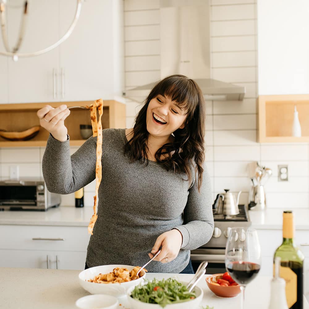 Jessie standing in a white kitchen twirling pasta with a fork and smiling.