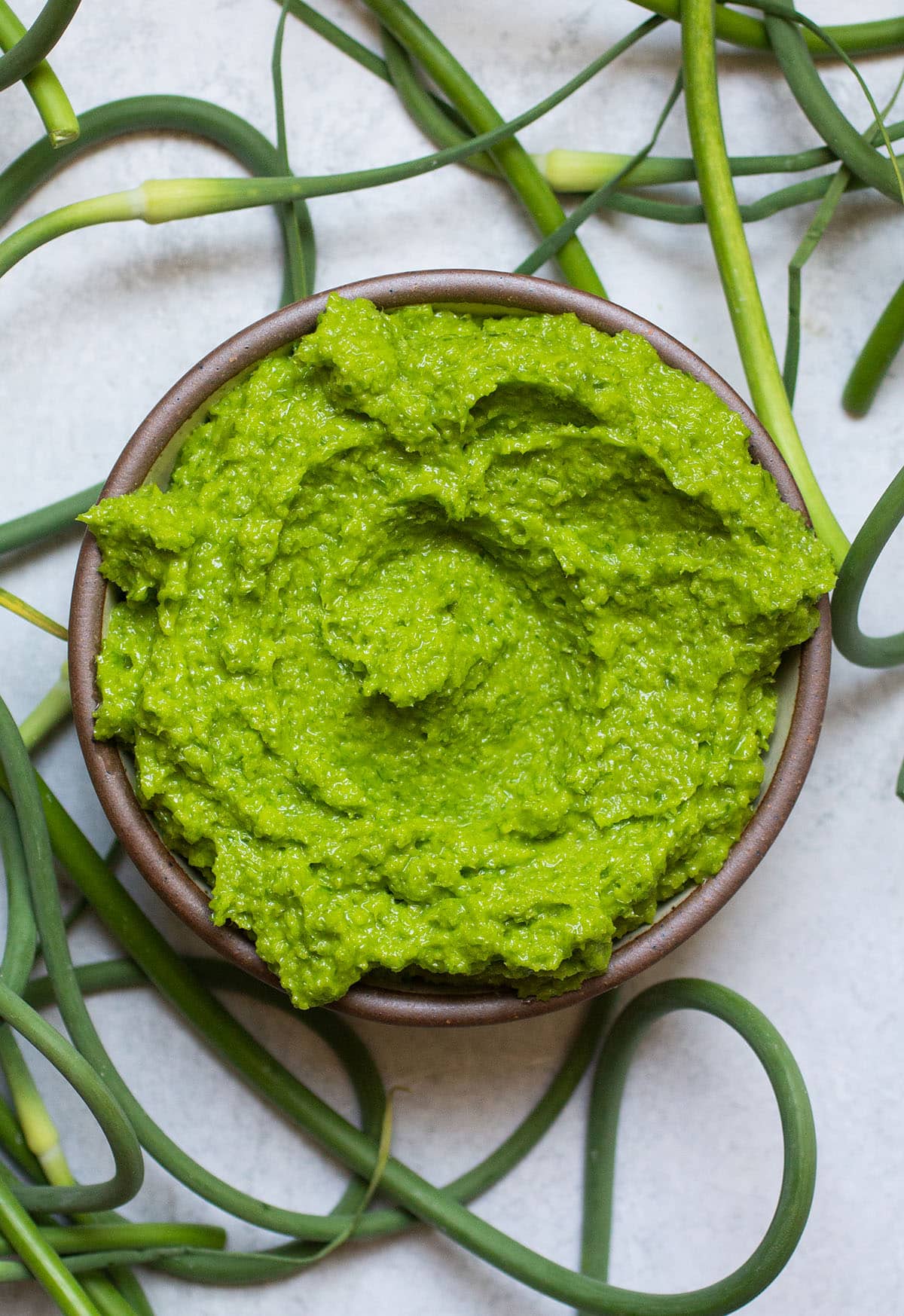 Garlic scape pesto on a white table surrounded by fresh garlic scapes.