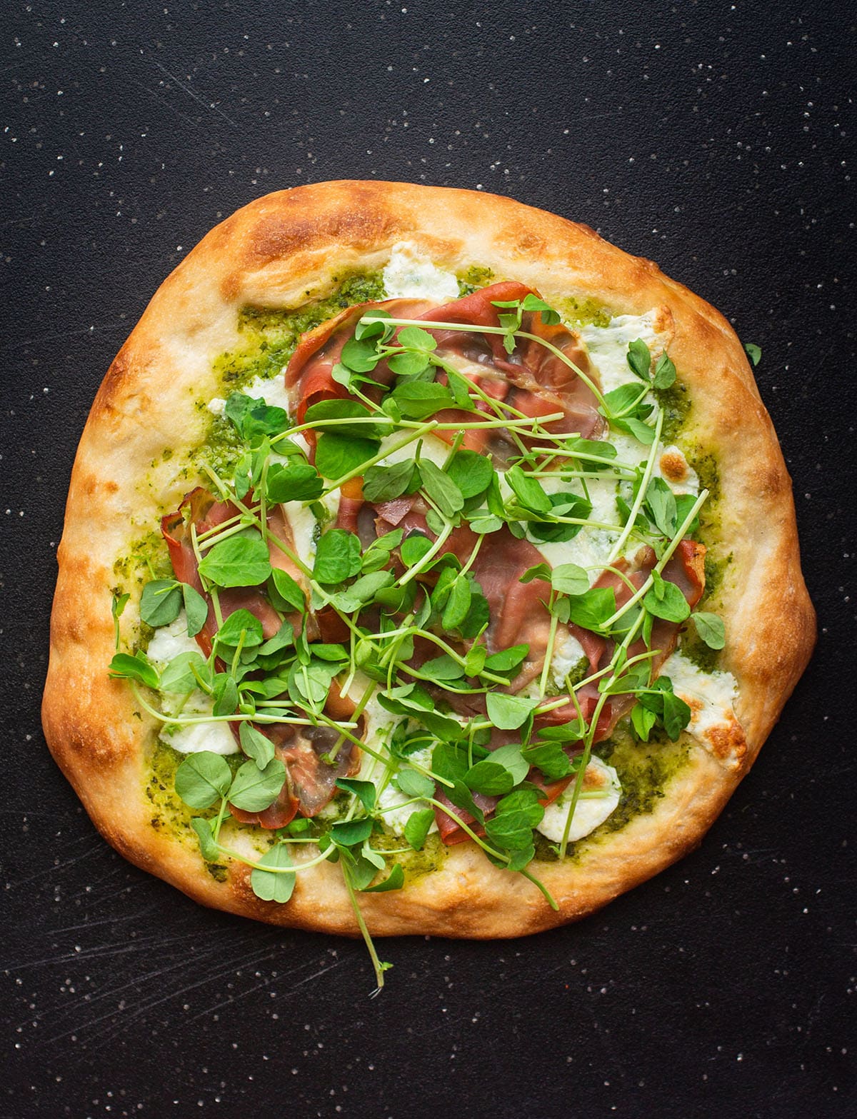 Pizza topped with fresh pea shoots on a black cutting board.