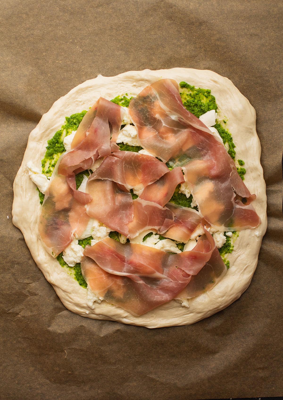 Pizza dough topped with pesto, cheese, and prosciutto on a piece of parchment paper.