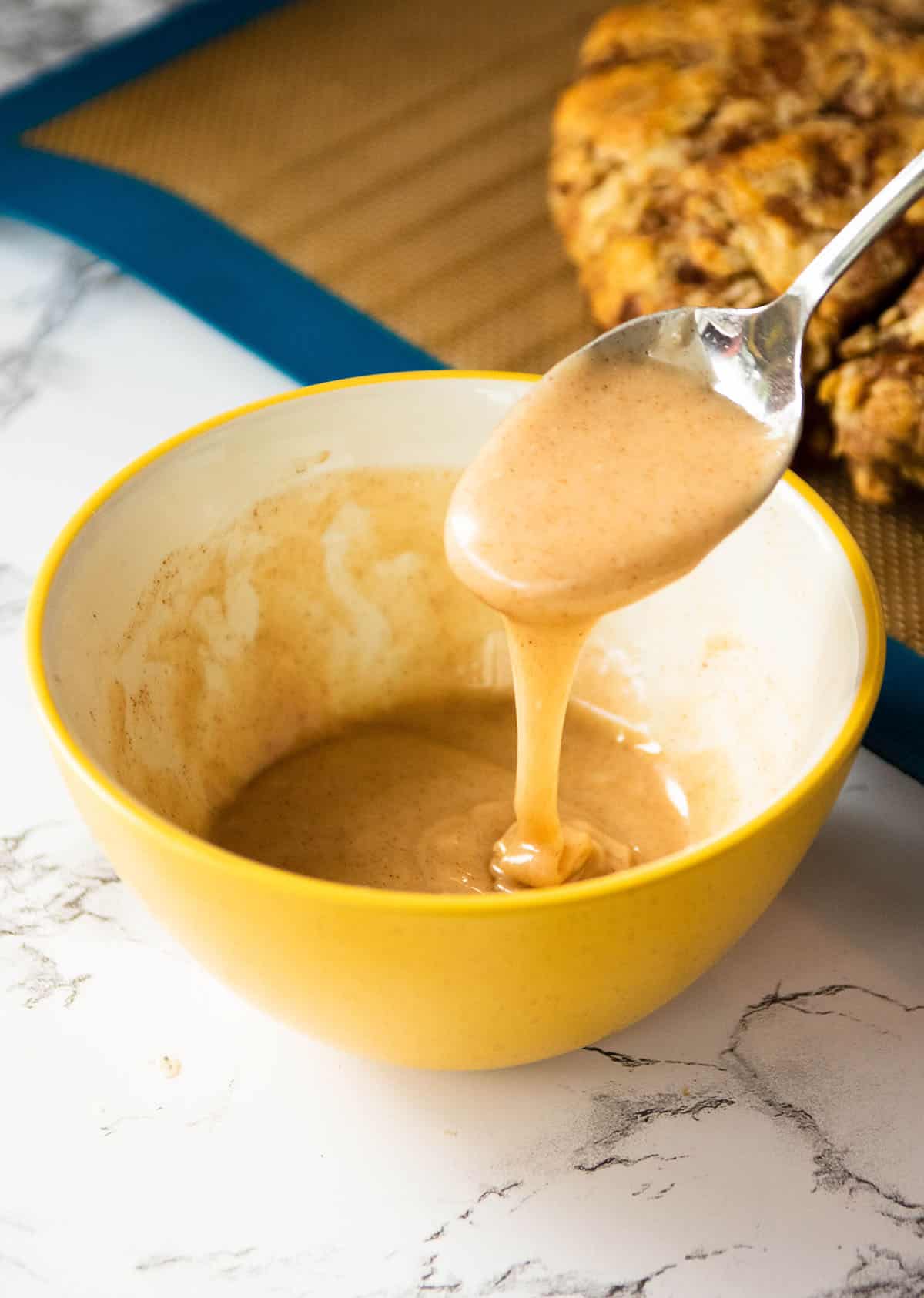 Spoon lifting a scoop of cinnamon glaze out of a yellow bowl.