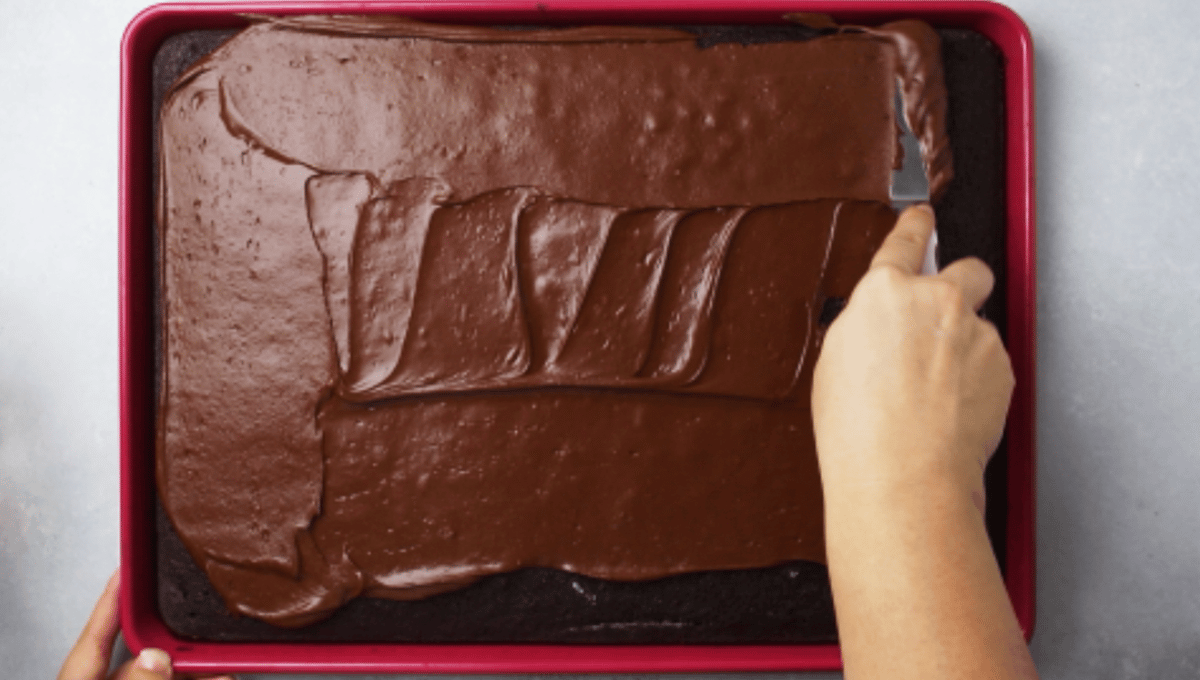 Hand using an offset spatula to spread frosting on a sheet cake.