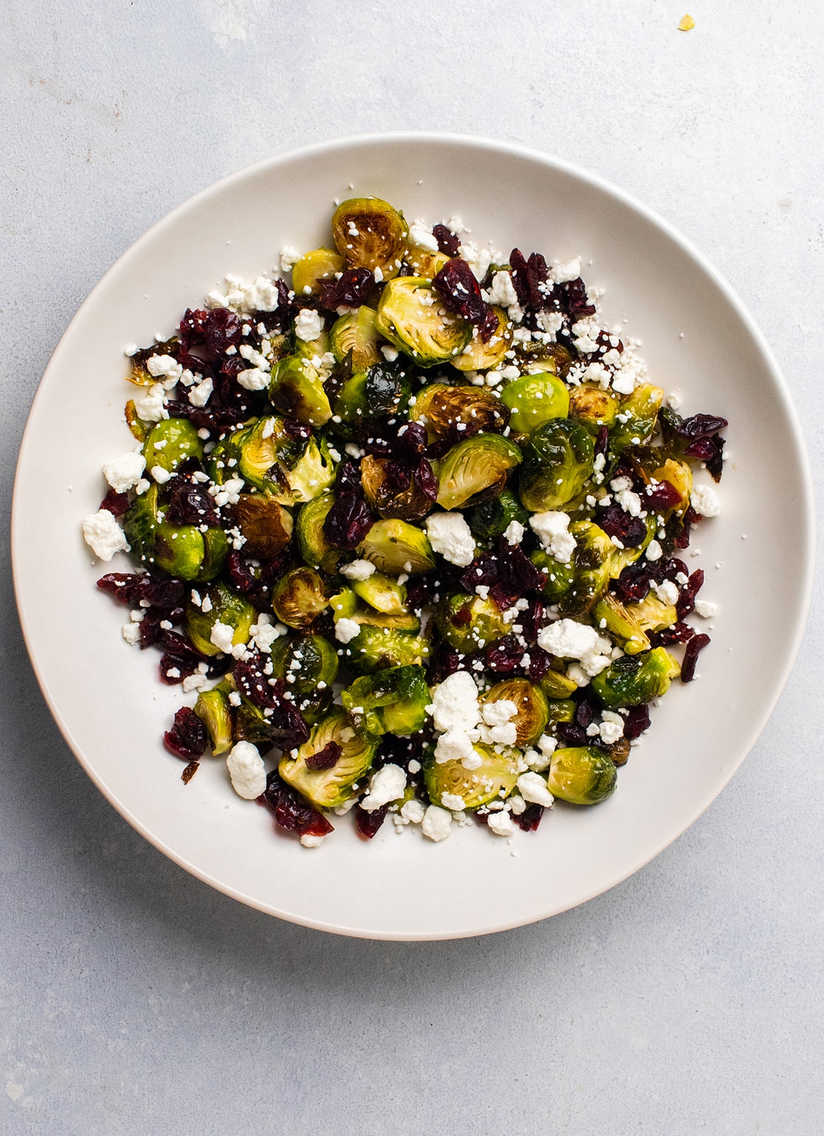 Overhead view of roasted brussels sprouts in a shallow white bowl with dried cranberries and crumbled goat cheese.