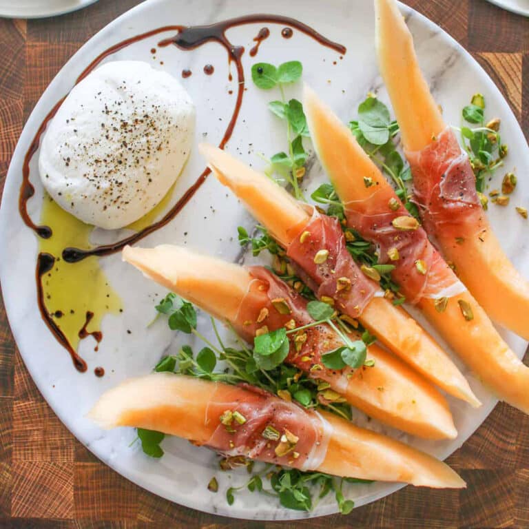 Melon slices wrapped in prosciutto on a white plate with pea tendrils and burrata.